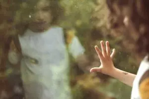 A child with their hand pressed to a window seeing their reflection.