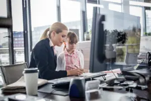 A working woman is visited by a child at her desk as they look at her computer. Horizontal shot.