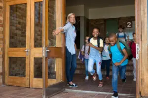 Smiling elementary school children happy to go home. Joyful school girls and boys wearing backpack and running outside the school at the end of the lessons. Teacher holding door while students running out.