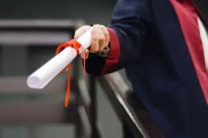 A diploma in a ribbon being help out by a hand wearing a graduation gown.