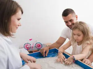 A therapist sitting at an activity center with a young girl and a father.