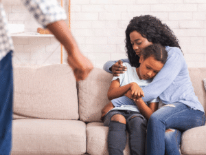Worried mother hugging sad daughter on couch while an angry adult figure stands in the foreground