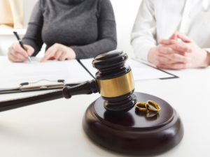 Close-up of a judge’s gavel and wedding rings on a desk with two individuals reviewing documents in the background, representing high-asset divorce proceedings and legal decision-making in San Diego County.