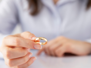 A person holds a gold wedding ring between their fingers, with a softly blurred background suggesting a moment of reflection or decision. The image symbolizes separation or the emotional transition of divorce, reinforcing the idea that each individual’s experience and outcome will be uniquely personal.
