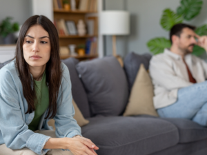 Woman sitting on a couch looking distressed while a man sits apart in the background, illustrating emotional strain and relationship conflict during legal separation or divorce decisions.