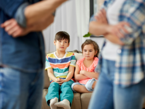 Two children sitting quietly while separated parents argue in the foreground, illustrating child custody disputes, co-parenting challenges, and decision-making for children with special needs.