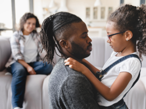Father holding a young child in a calm home setting while another parent sits nearby, illustrating family relationships and the child-focused considerations involved in custody decisions.